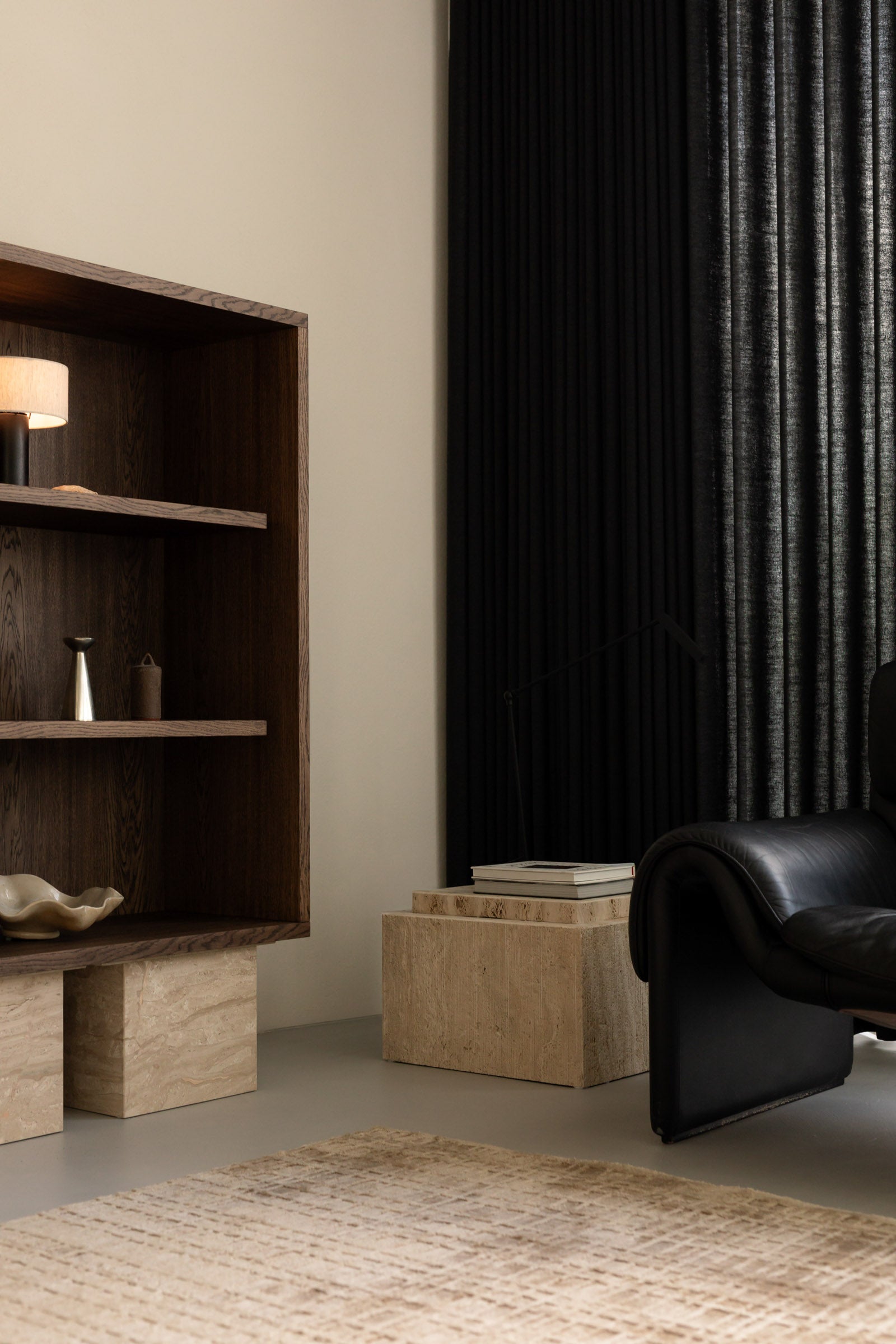 Un'common dark oak BLOCK Cabinet shelving unit with a table lamp and ceramic pieces, paired with travertine BARI side tables and a black leather lounge chair against a pleated black curtain.