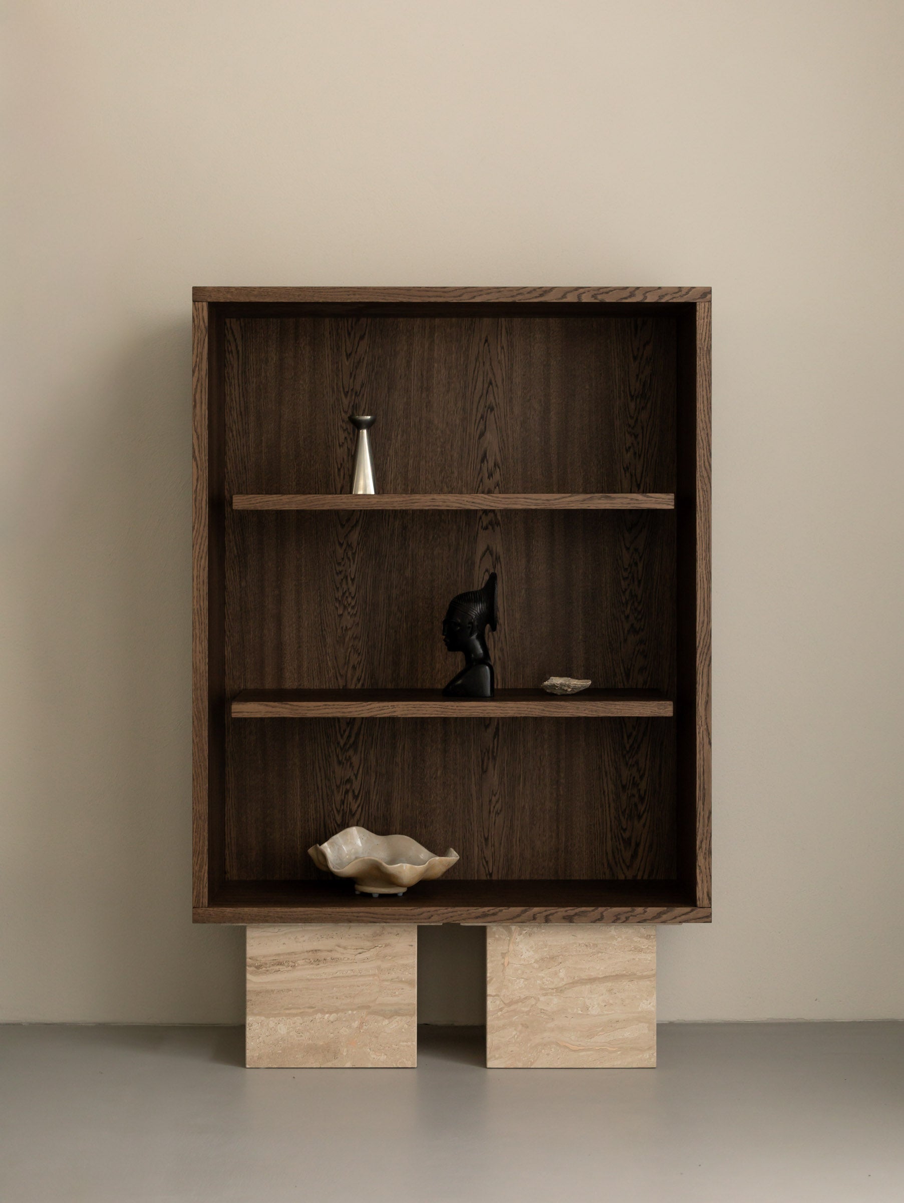 Un'common BLOCK cabinet in dark oak on travertine block legs, styled with a brushed metal vase, a carved bust sculpture, a small stone, and a scalloped ceramic bowl