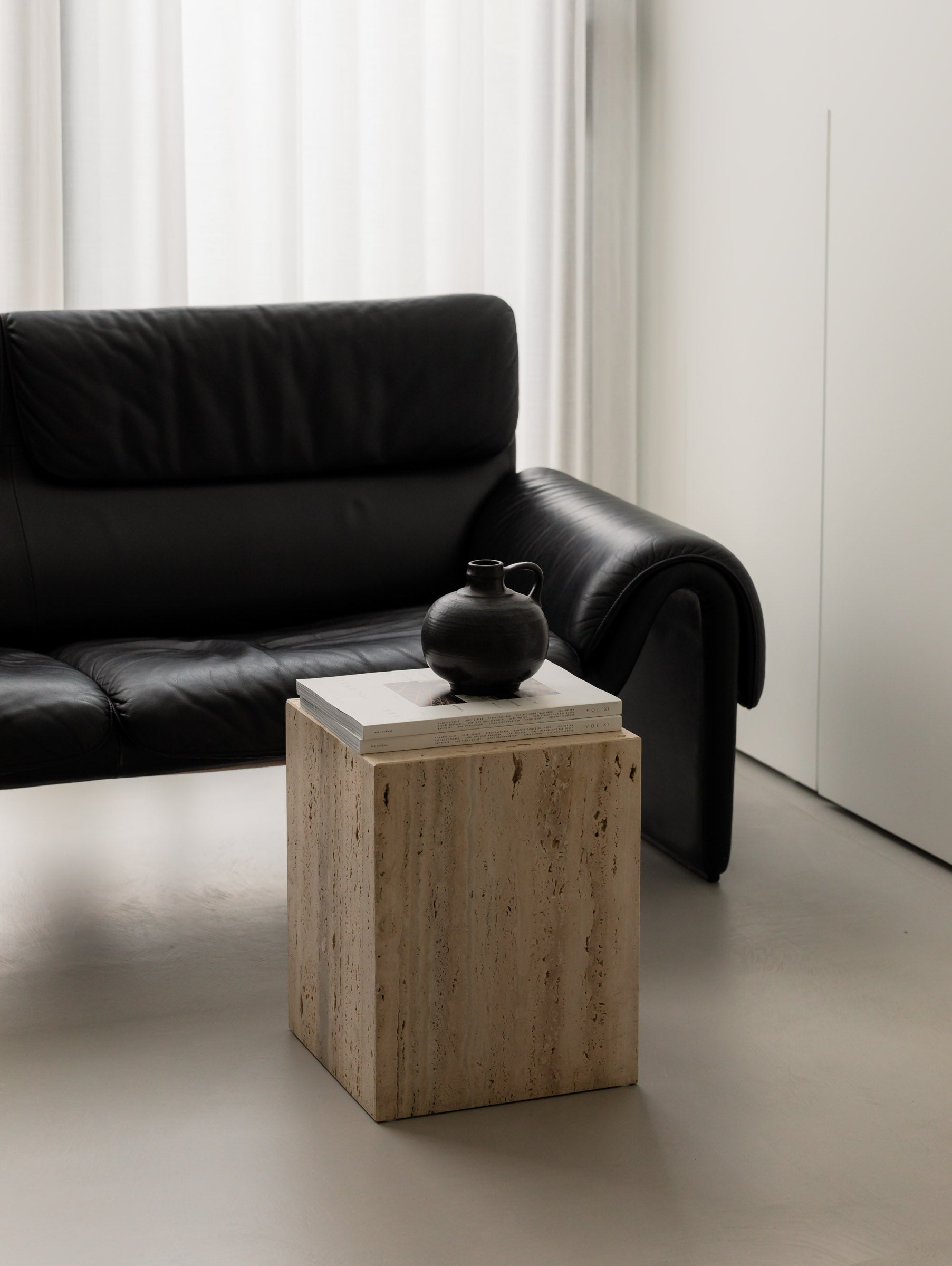 Un'common COI travertine pedestal with a round black ceramic jug on stacked art books, in front of a black leather lounge chair and sheer curtains.